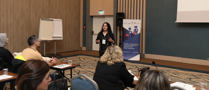 Betimleme: Konferans salonunda Ayşe Sarı ayakta konuşma yapıyor. Katılımcılar masa başında oturuyor, önlerinde su bardakları, mikrofonlar ve belgeler var. Sarı'nın arkasında bir yazı tahtası ve dikey bir afiş bulunuyor. Afişte ILO, Engelsiz Kariyer logoları yer alıyor ve “Engelliler ve İşverenler için Farkındalık Eğitimleri ve Engelsiz Kariyer Günleri Buluşmaları” yazıyor. Salon sade ve profesyonel görünüyor.